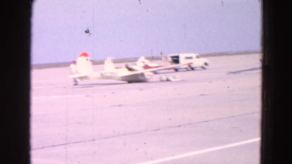 A small white aircraft is seen from the rear on a bright 1964 airfield, with another aircraft and a van in the distance.