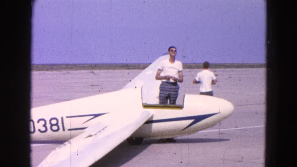 A man stands in the cockpit opening of a white glider marked 0381 during apparent preflight activity at the airfield.