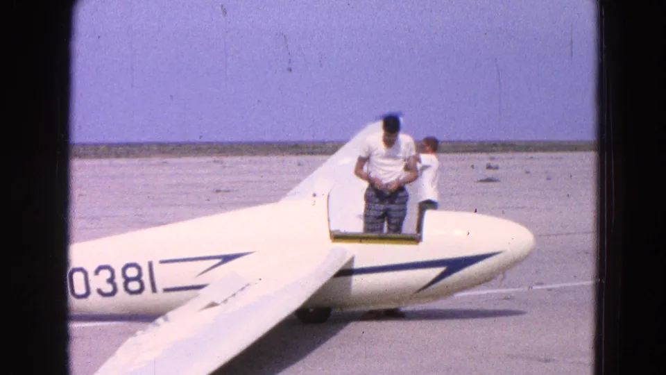 A man leans into the cockpit area of a white glider as another person stands nearby on the sunlit tarmac.