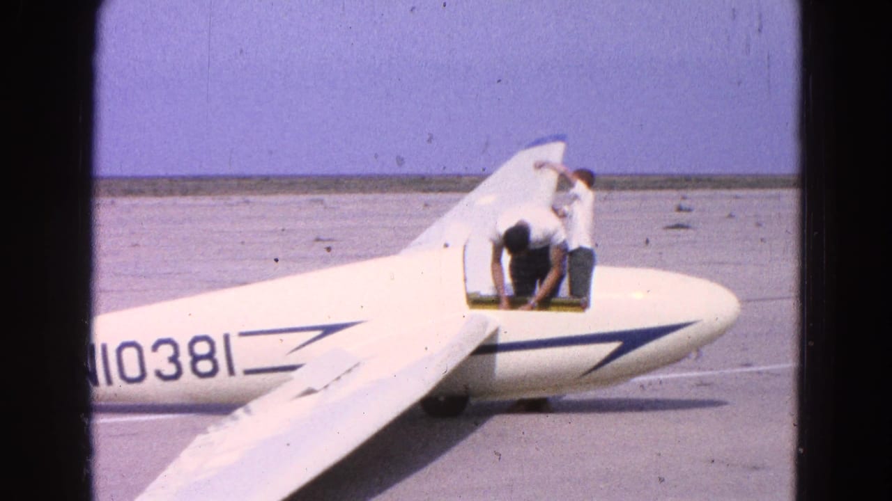 Two men inspect a white glider on an airfield beside a partial aircraft number.