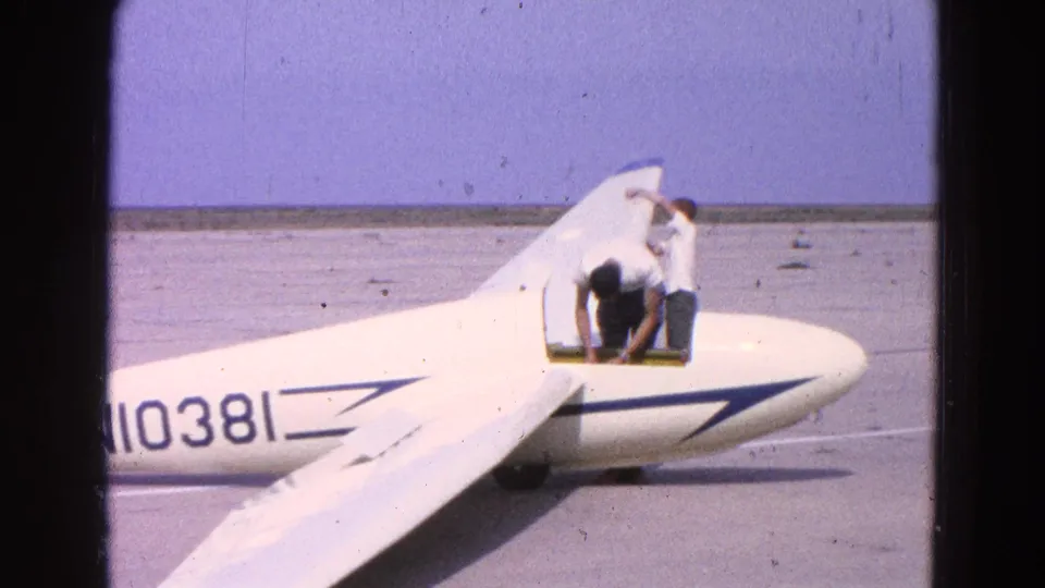 1964 home-movie frame of two men at a white glider on a Washington DC airfield, with blue markings and partial number visible.