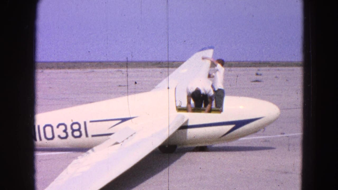 The white glider sits on the airfield as two men continue inspecting or entering the cockpit area in this 1964 frame.
