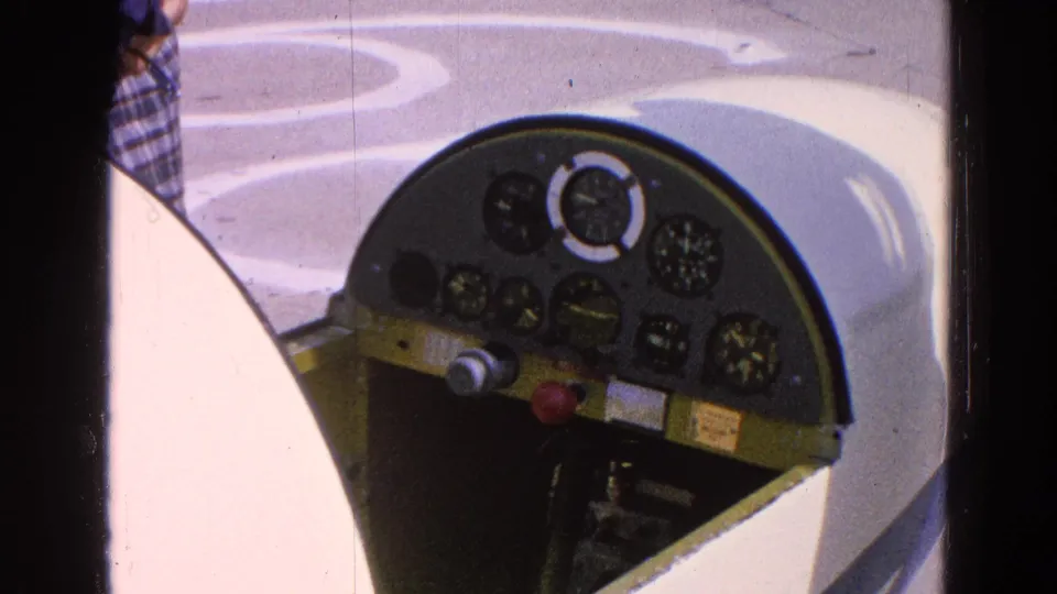Side-angle cockpit detail from the 1964 Washington, DC clip, with analog gauges, pale aircraft bodywork, and a partially visible person nearby.