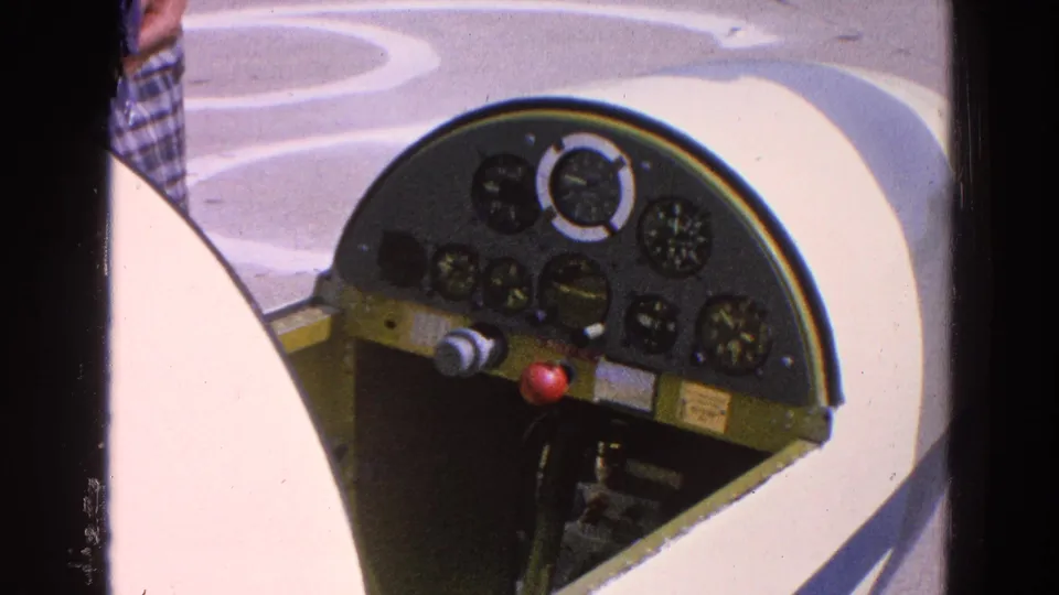 The cockpit remains in close view, showing round gauges, a red knob, light aircraft surfaces, and a cropped figure in plaid clothing.