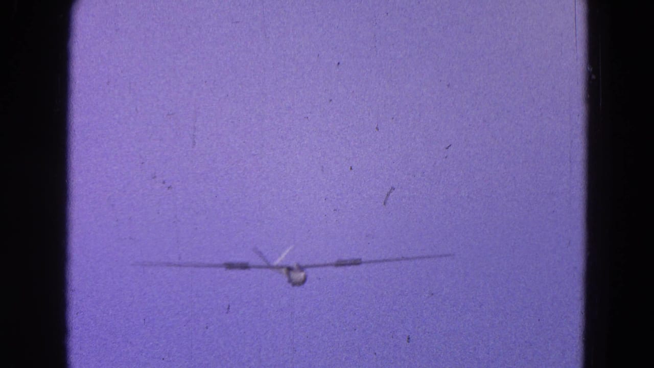1964 Washington, DC home-movie frame showing a distant small propeller plane head-on in a purple-tinted sky.