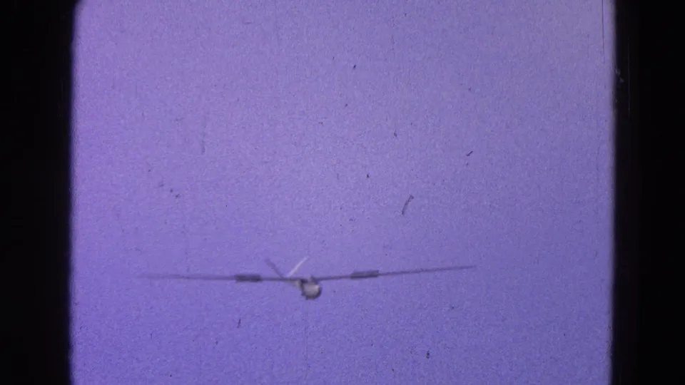 1964 Washington, DC home-movie frame showing a distant small propeller plane head-on in a purple-tinted sky.