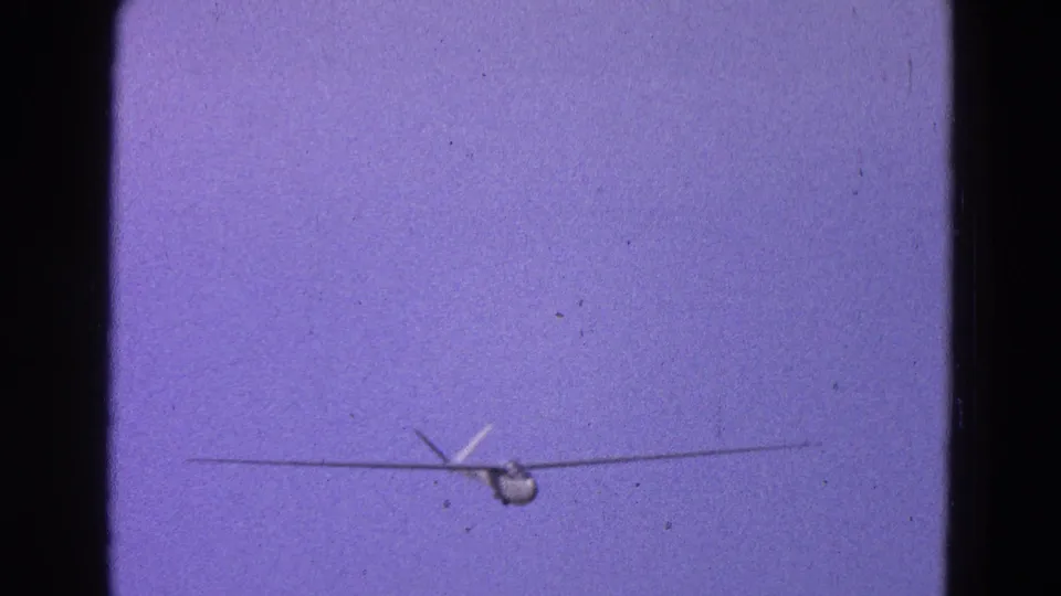 Home-movie still of a small propeller plane lower in the frame, with soft focus and purple-shifted 8mm color.