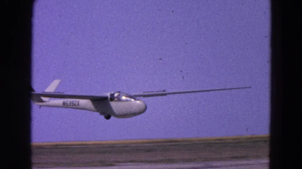 The glider passes low over the runway, showing its canopy, long wing, tail, and partial registration marking.