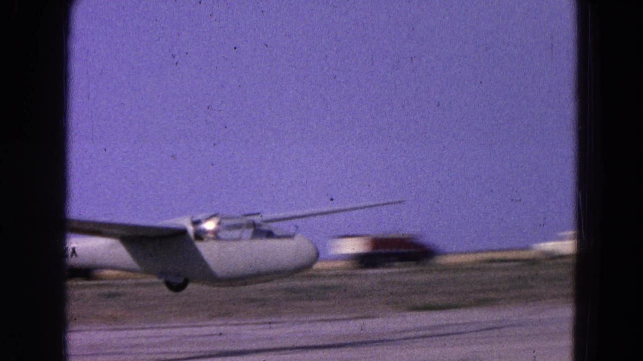 White glider in a 1964 Washington, DC area home-movie frame, moving low across a runway with distant airfield buildings.
