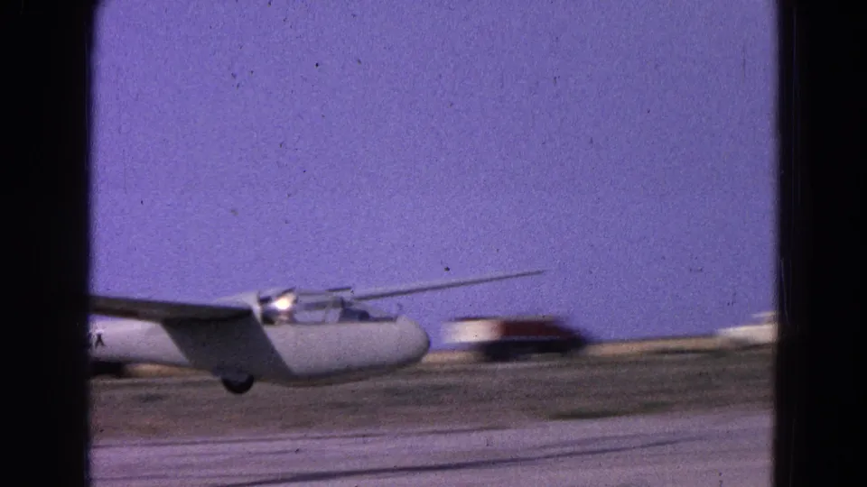 White glider in a 1964 Washington, DC area home-movie frame, moving low across a runway with distant airfield buildings.