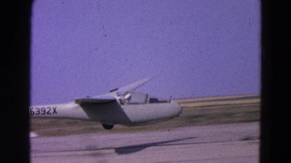 White glider lifting off from a runway in 1964 8mm footage, with its shadow trailing below.