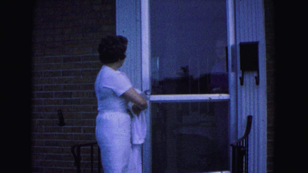 Woman in a white dress stands at a brick house doorway holding a garment.