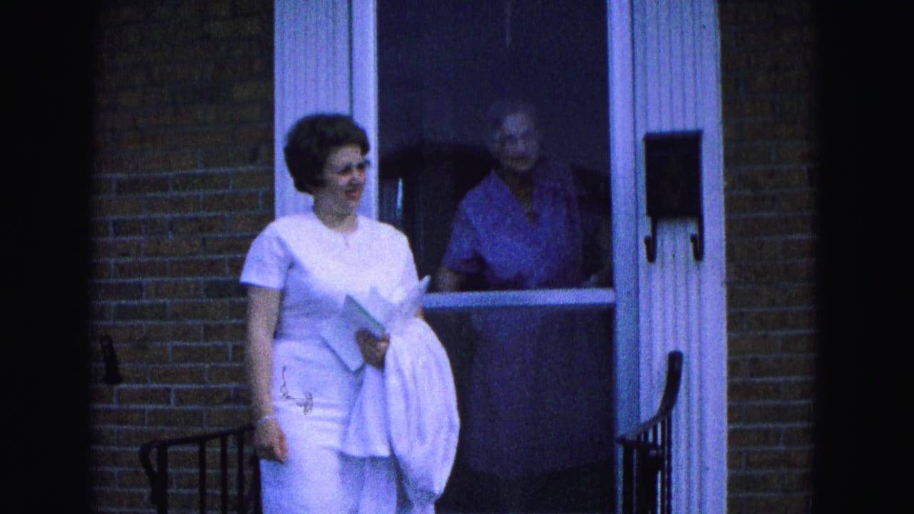 Woman steps outside a brick home while an older woman is visible behind the screen door.