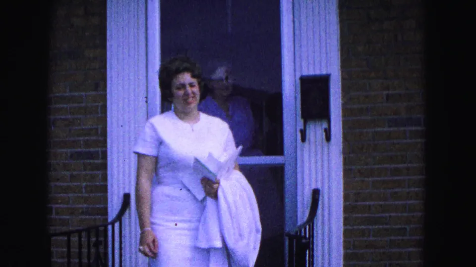 The woman walks away from the doorway of the Williamsburg home, carrying papers or a book and a white garment.