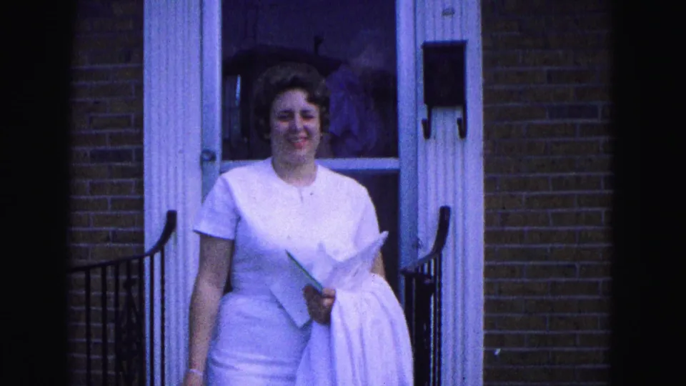 A smiling woman in white faces the camera outside the 1964 Williamsburg brick home, with another woman faintly visible inside.