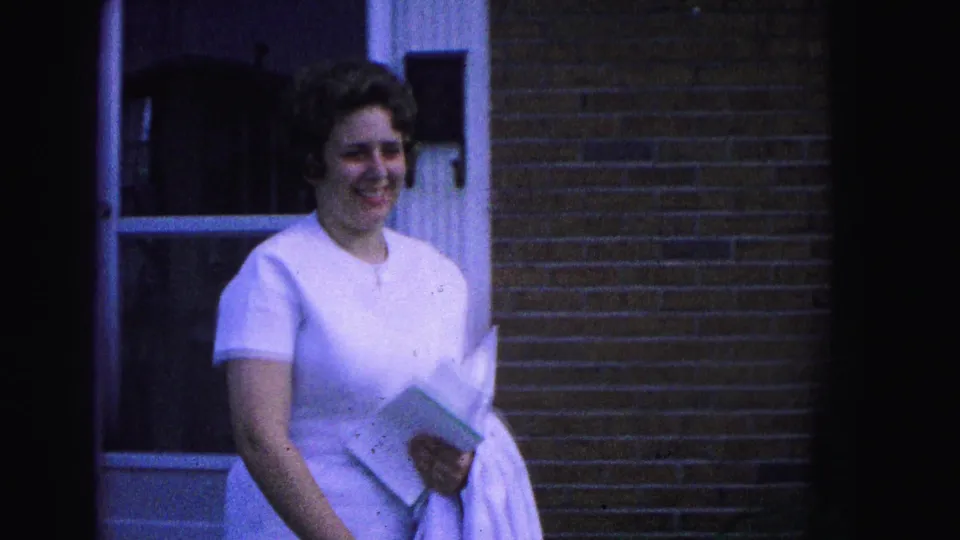 In Williamsburg, Virginia home-movie footage from 1964, a woman in a white dress stands near a brick house entrance holding papers.