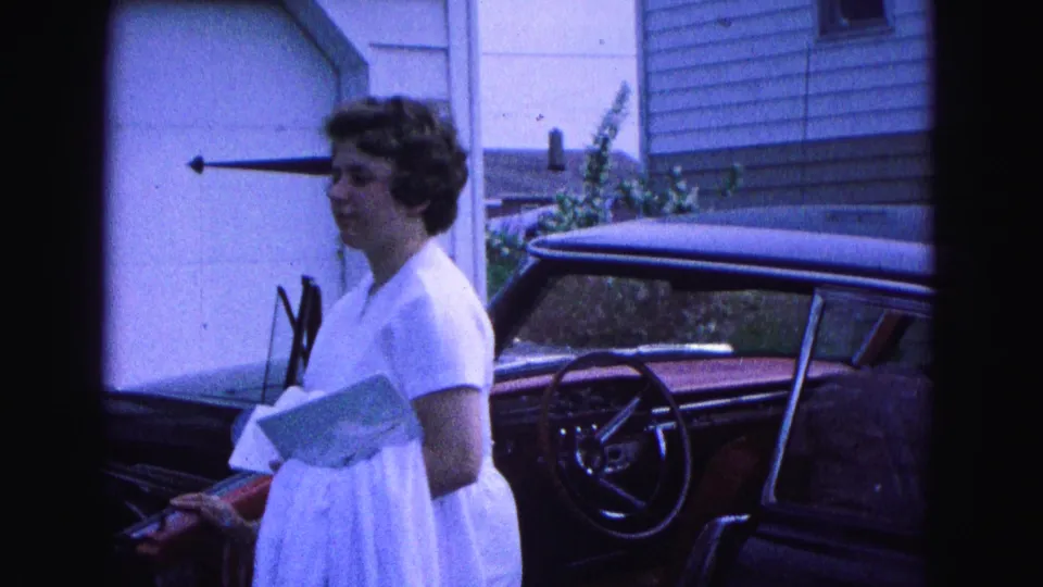 A woman in a white dress walks beside a dark vintage car outside a residential building in Williamsburg, Virginia, 1964.