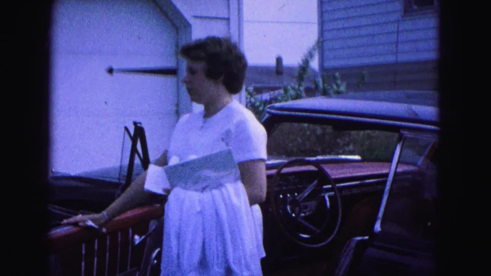 The woman pauses by a dark car with the door open, holding papers outside a Williamsburg residence in 1964 home-movie footage.