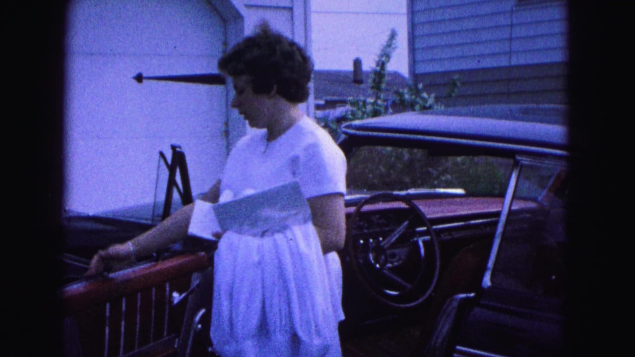 Woman in a white dress stands beside a red classic car holding paper near a garage.