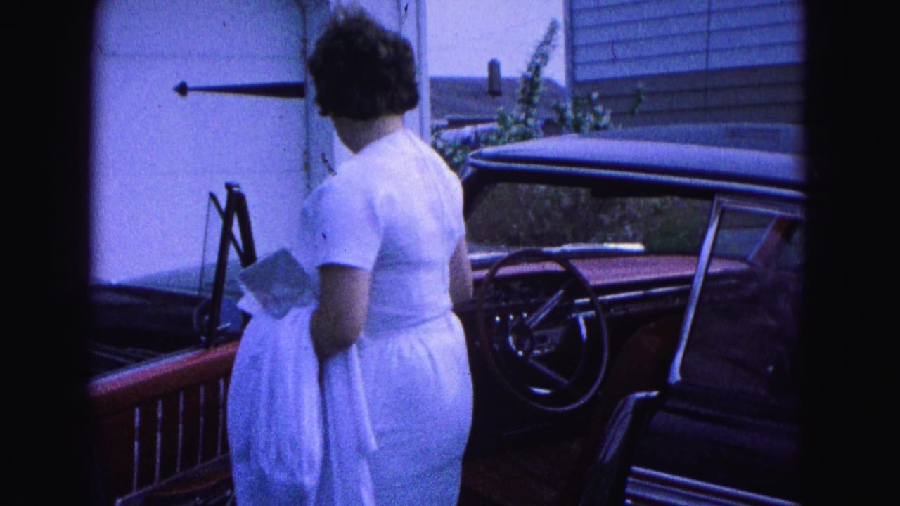 Woman turns toward the open driver's door of a red classic car outside a garage.