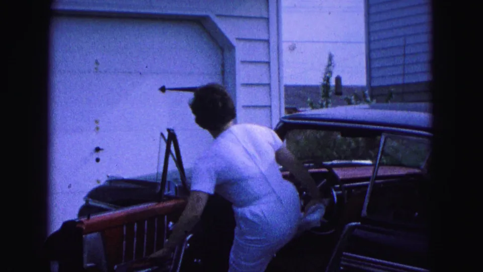 The woman leans into the red classic car as she prepares to enter, with the garage door and vehicle interior visible in the frame.