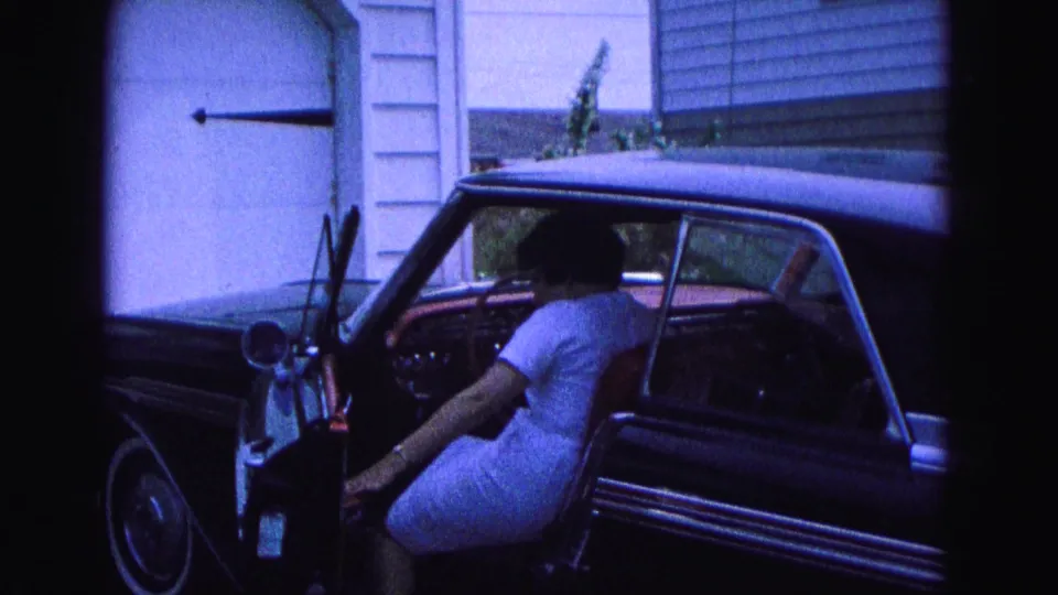 The woman is seated in the driver's area of the red classic car, with the open door, steering wheel, dashboard, and residential exterior visible.