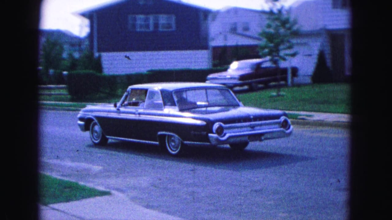 Dark vintage sedan driving on a suburban street past houses and lawns.