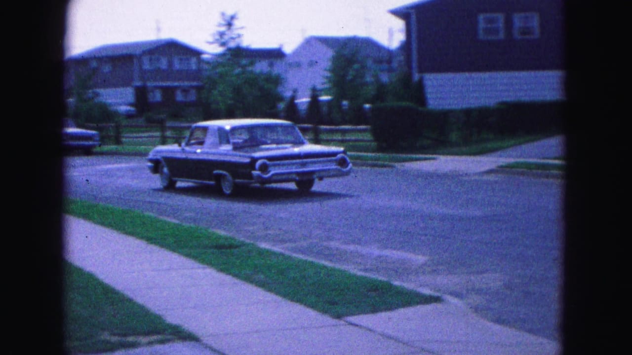 The dark sedan continues through a Williamsburg residential street scene, with sidewalks, lawns, and midcentury houses visible.