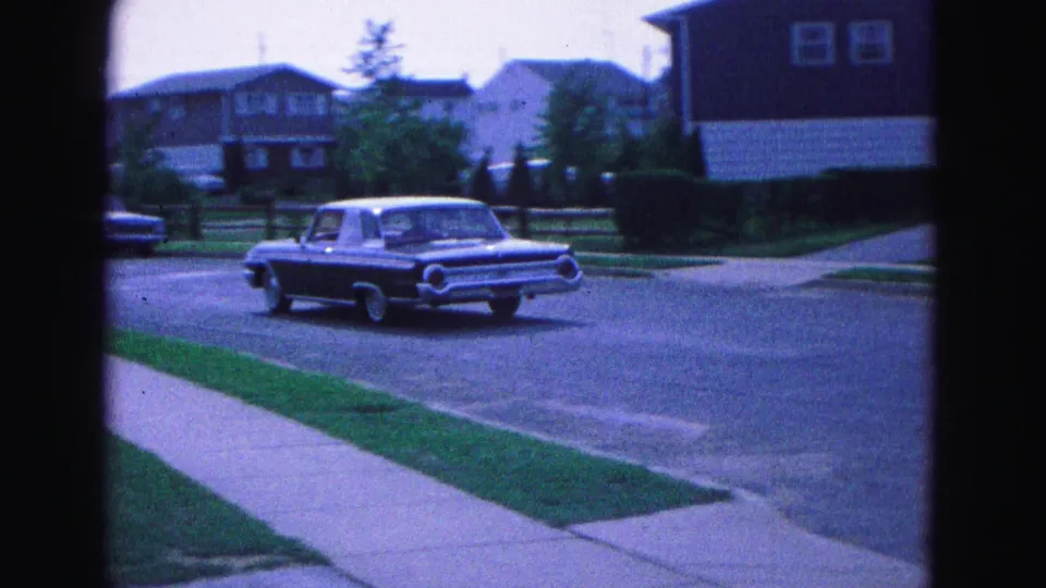 The dark sedan continues through a Williamsburg residential street scene, with sidewalks, lawns, and midcentury houses visible.