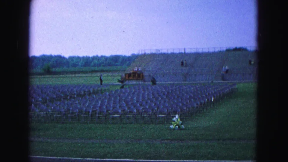 Rows of folding chairs fill a grassy outdoor field in Williamsburg, with a small platform area and distant people visible in 1964 footage.