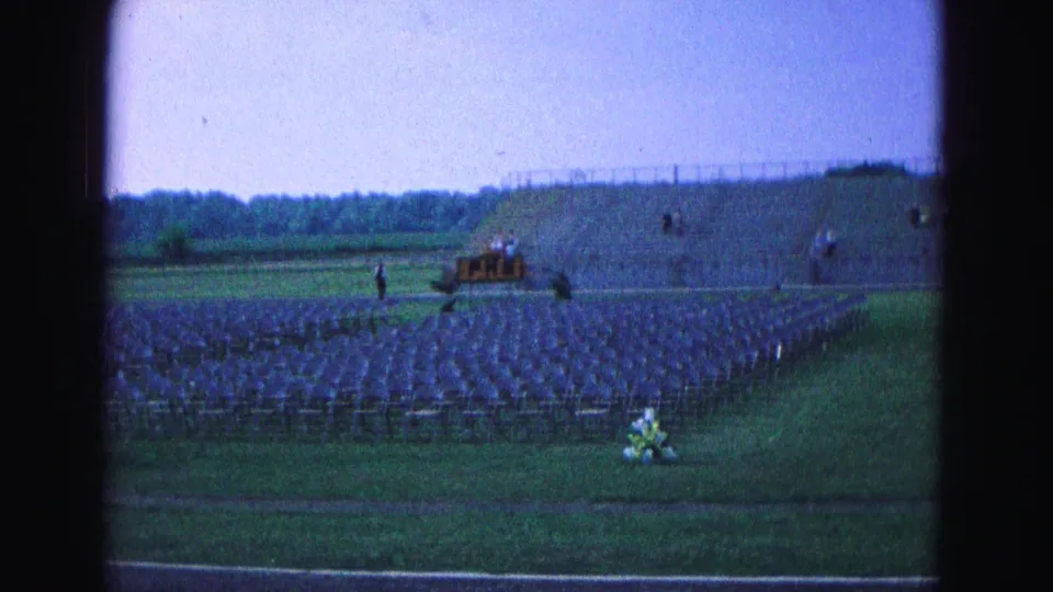 A wide home-movie view shows organized folding chairs on a grassy field in Williamsburg, with distant figures near a platform area.