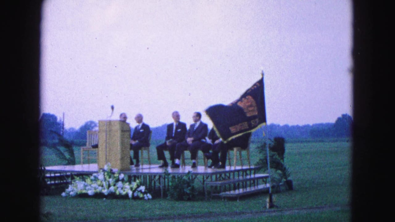 Men in suits sit on an outdoor stage beside a podium and school flag.