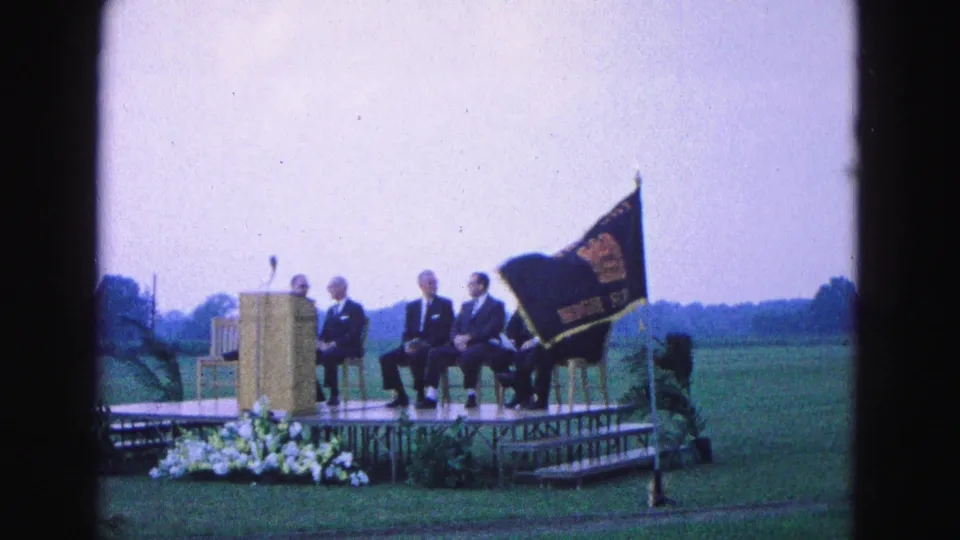1964 Williamsburg outdoor ceremony view with suited men seated near a podium and a dark school flag on a raised platform.