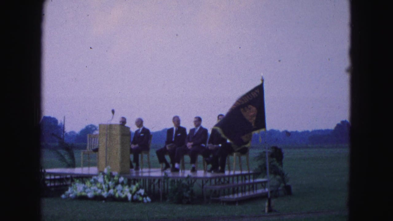 Williamsburg, Virginia ceremony frame showing seated men, a podium with microphone, and a dark school flag on a grassy field.