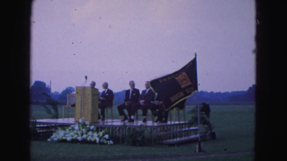 1964 home-movie frame of a dark school flag moving across a stage where suited men sit beside a podium in Williamsburg.