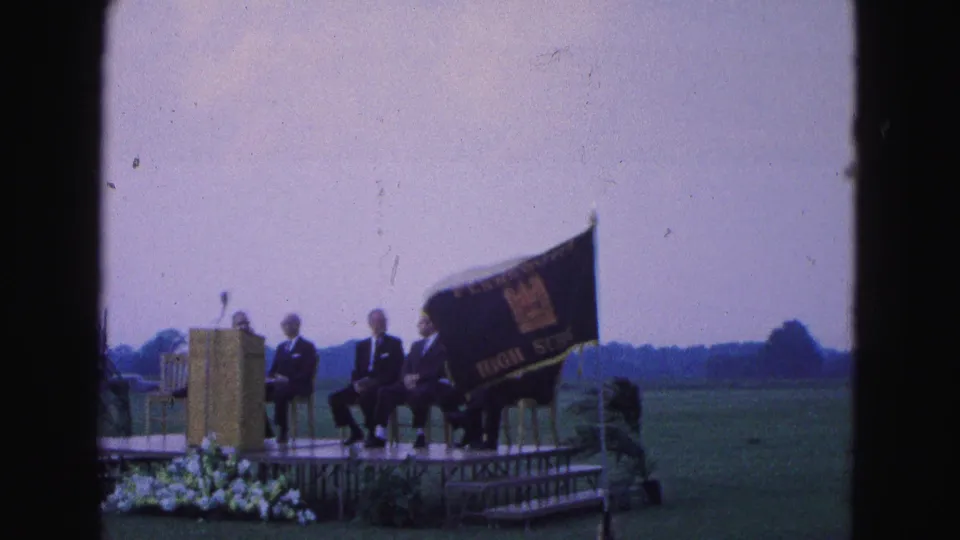 Williamsburg school ceremony footage with a large dark flag in front of seated men, a podium, flowers, and an open field.