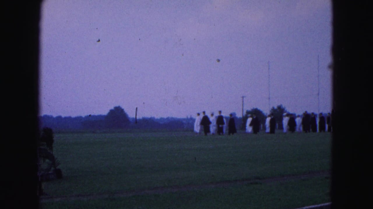 Graduates in dark and light robes stand in a distant line across a grassy field.