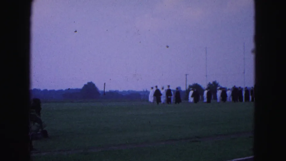 1964 Williamsburg, Virginia home-movie frame showing a distant graduation class lined up on grass under a pale sky.