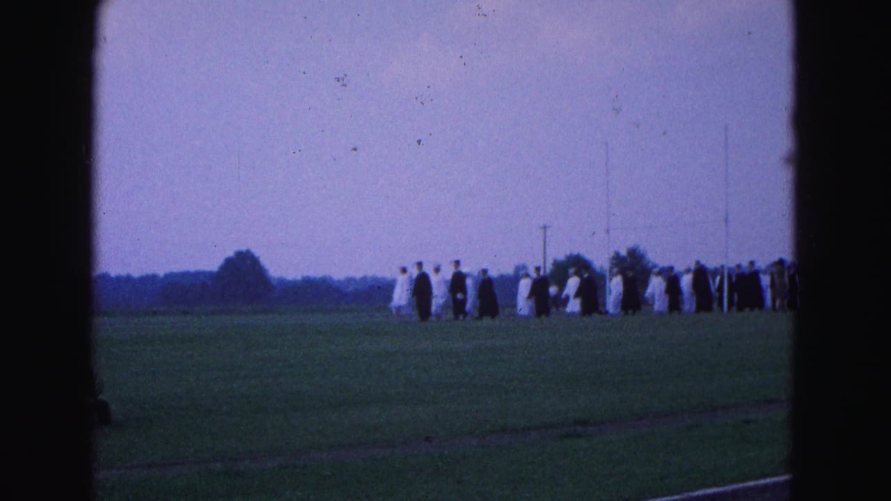 A row of graduates in robes extends across the field near distant trees and poles.