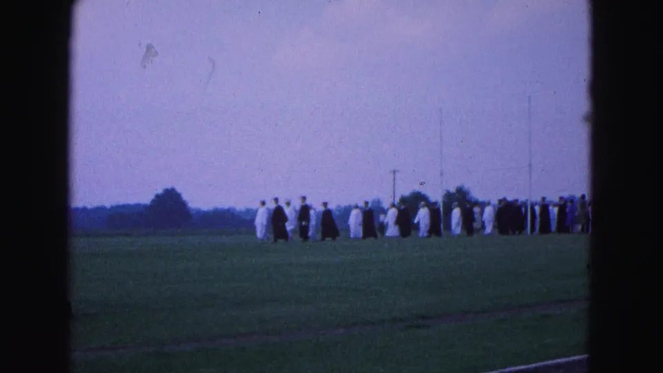 Distant 1964 commencement scene with robed graduates arranged in a long line on grass in Williamsburg, Virginia.
