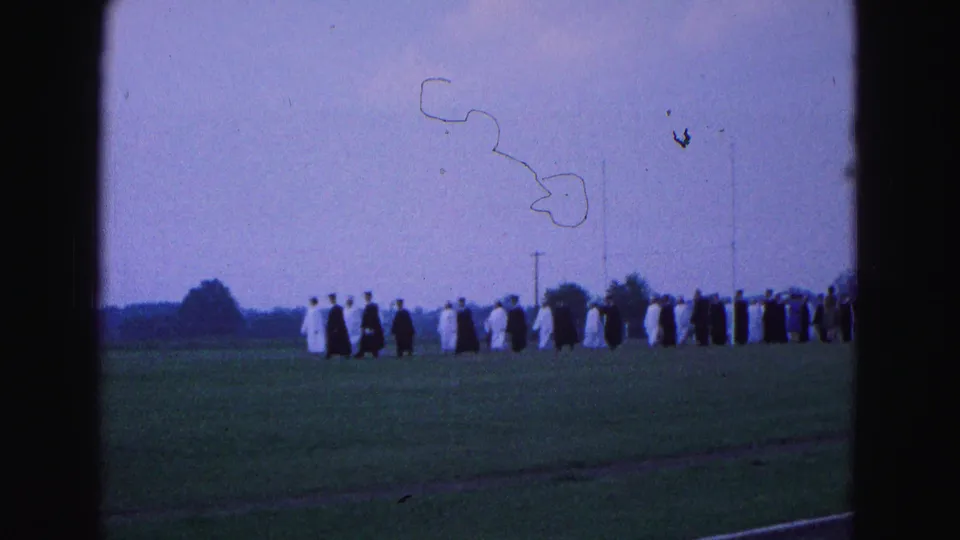 Wide home-movie frame of robed graduates on a Williamsburg field, with visible dust and frame marks from the 8mm scan.