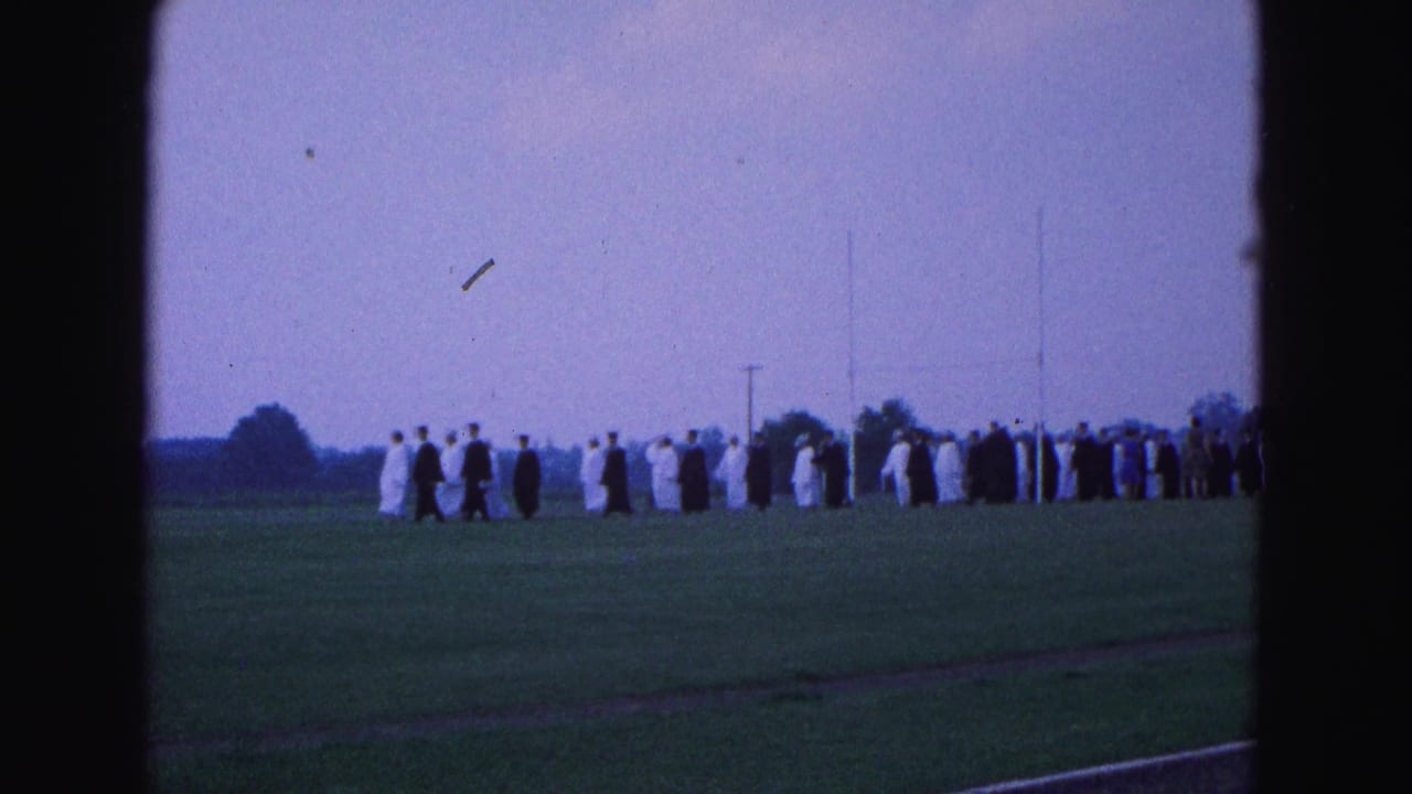 Graduates in black and white robes walk across a grassy field.