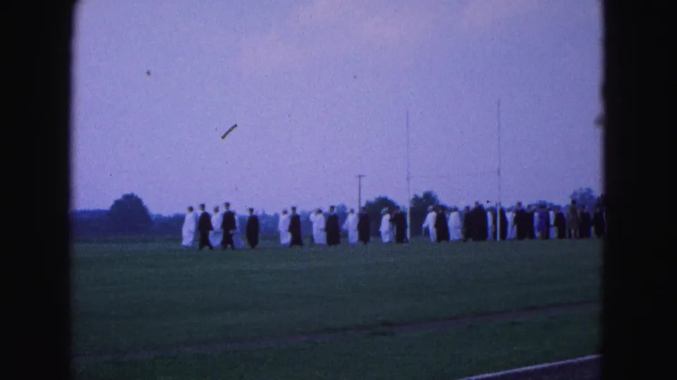 In 1964 Williamsburg, Virginia footage, robed graduates process in a line across an open grassy field.