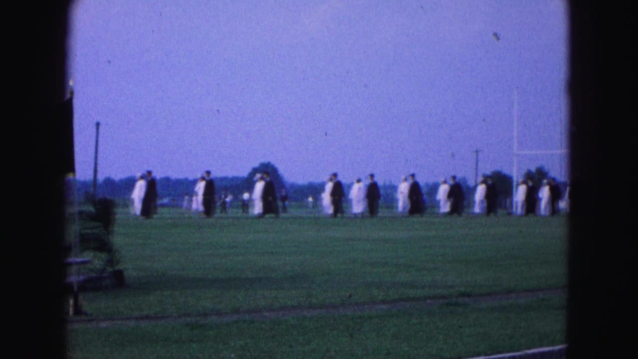 Graduates in caps and gowns walk in a long line across a grassy field.