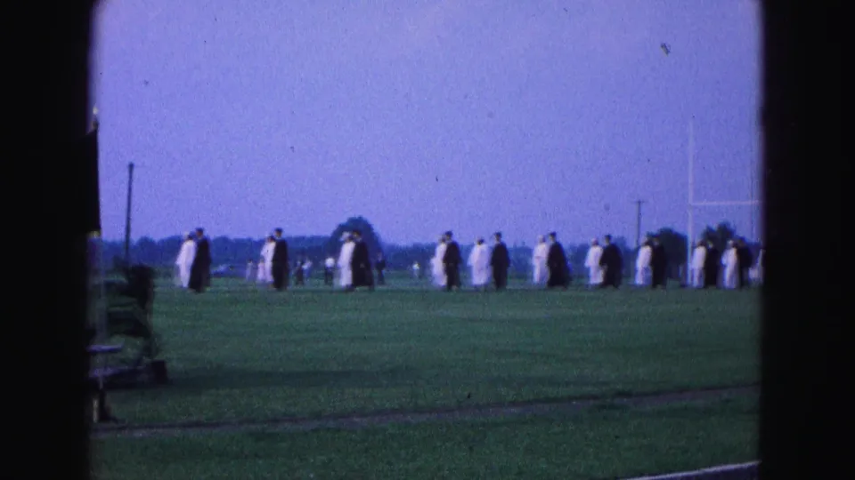 1964 Williamsburg, Virginia home-movie frame showing graduates in caps and gowns crossing a field with goalposts in the distance.