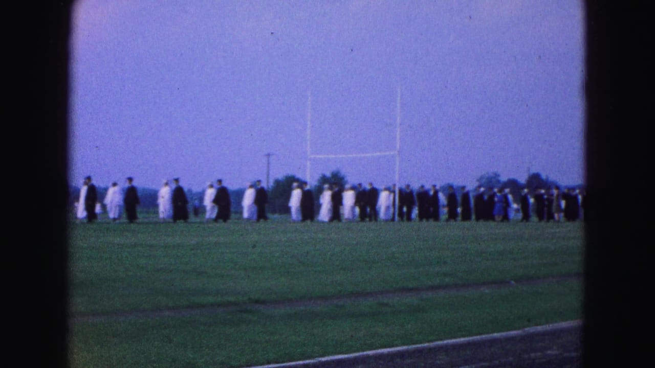 A long line of robed graduates continues across the field near distant goalposts.