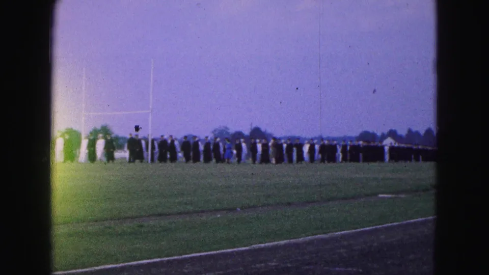 A distant 1964 graduation procession in Williamsburg shows alternating dark and light gowns crossing the athletic field.