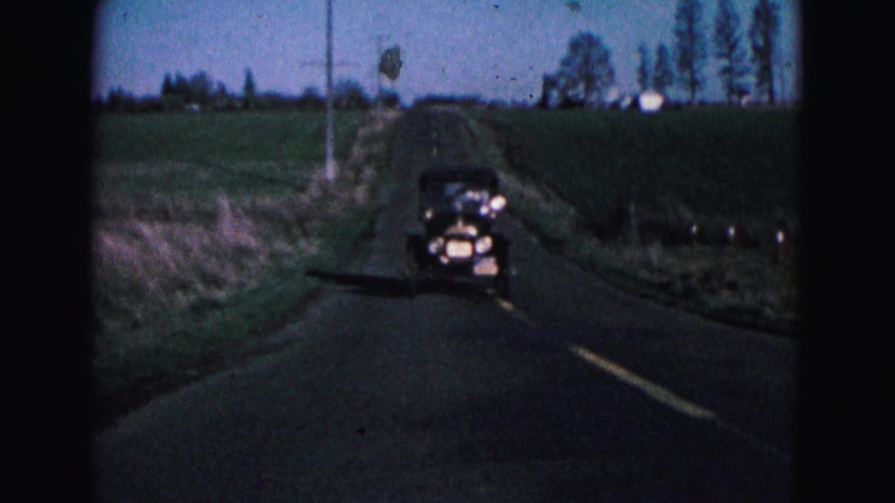 Antique black car approaches on a rural two-lane road bordered by fields.