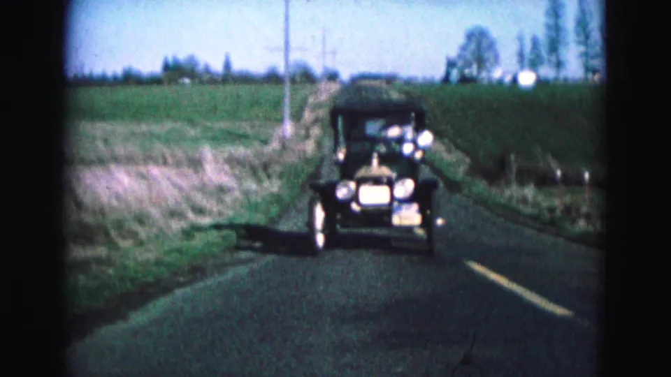The antique car nears the camera on a rural Williamsburg road, with fields, poles, and trees visible behind it.