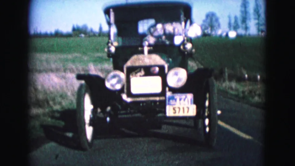 A close frame shows the front of the antique black car, including round lamps, radiator, windshield, and license plate details.